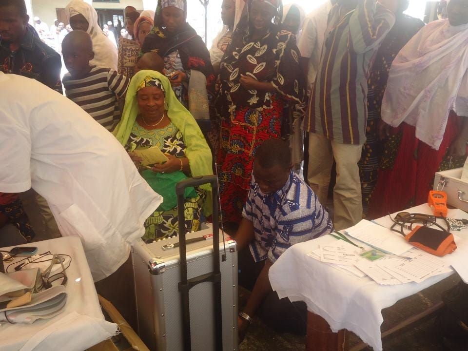An older woman smiling as she waits for her check-up, with a queue of people behind her
